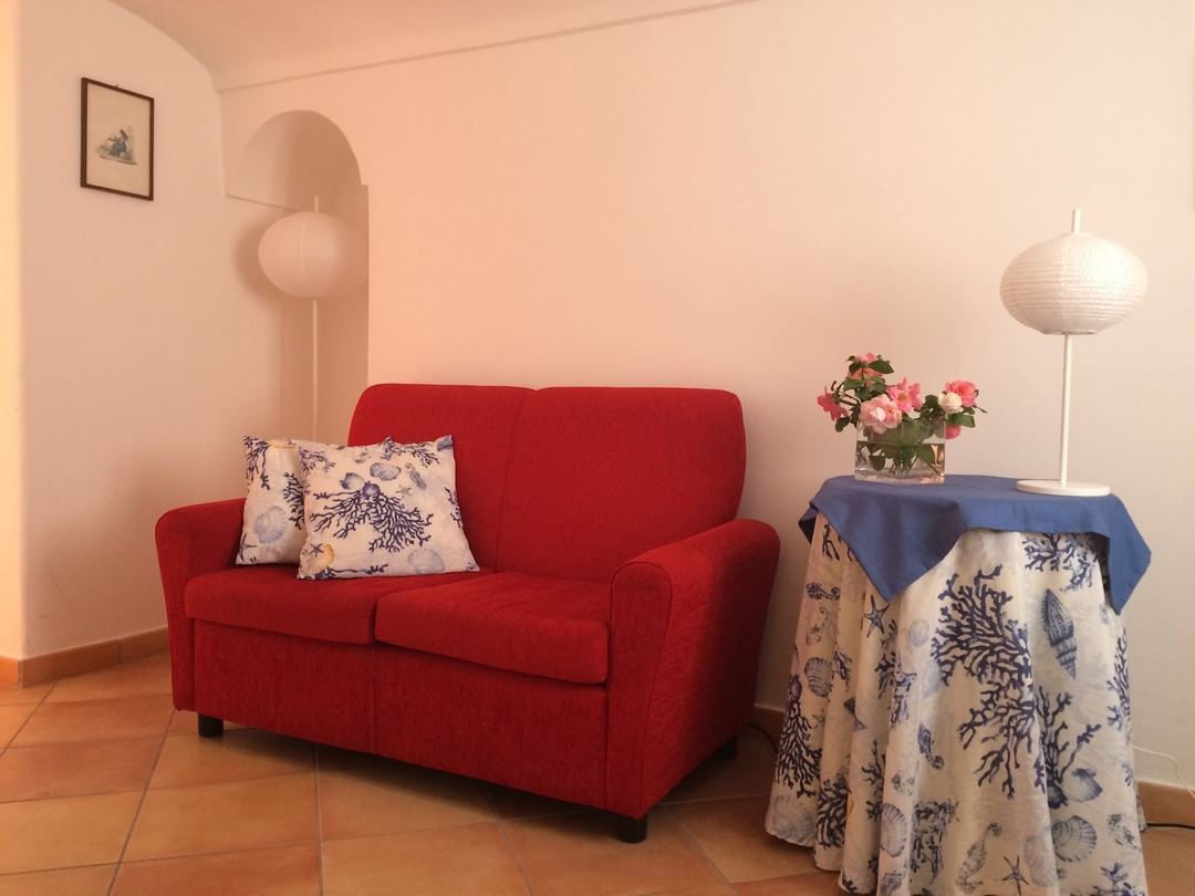 A bright, minimalist room interior featuring a red upholstered sofa with decorative pillows, a side table with fresh pink flowers, and soft ambient lighting.