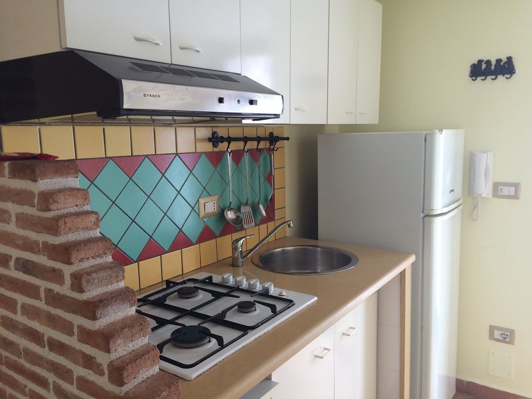 A compact kitchen featuring a gas stovetop, colorful turquoise and burgundy diamond-patterned backsplash, range hood, and white cabinetry.
