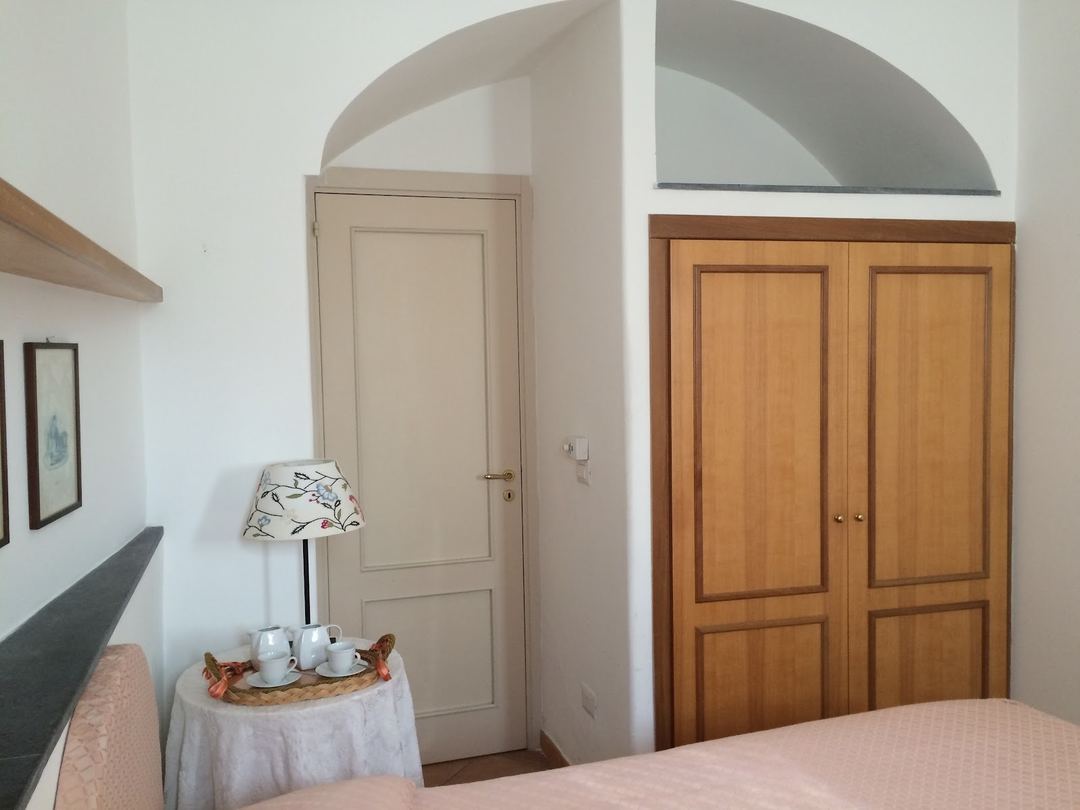 A bedroom corner featuring a wooden wardrobe, cream-colored door, small table with lamp and tea service, and arched architectural details.