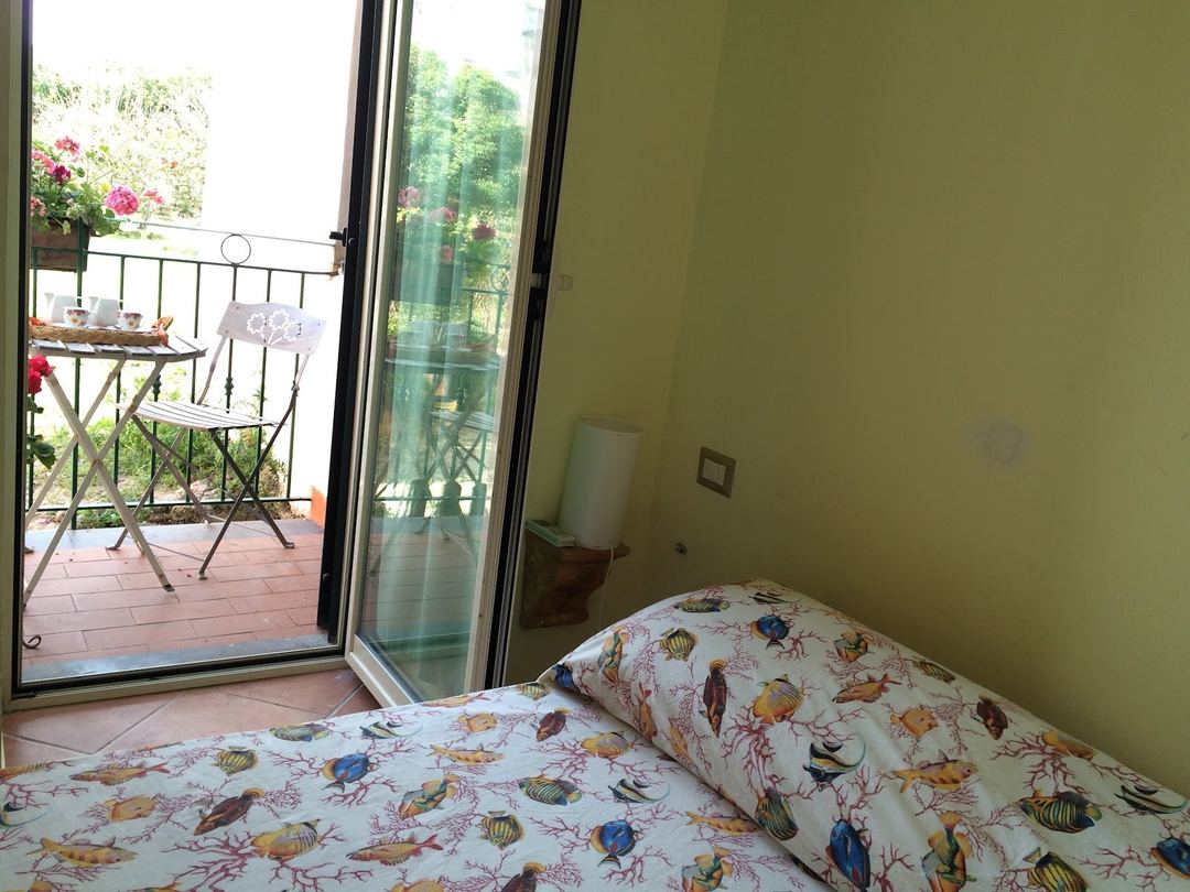 A bright bedroom with fish-patterned bedding opens onto a terrace with flowering plants and outdoor seating.
