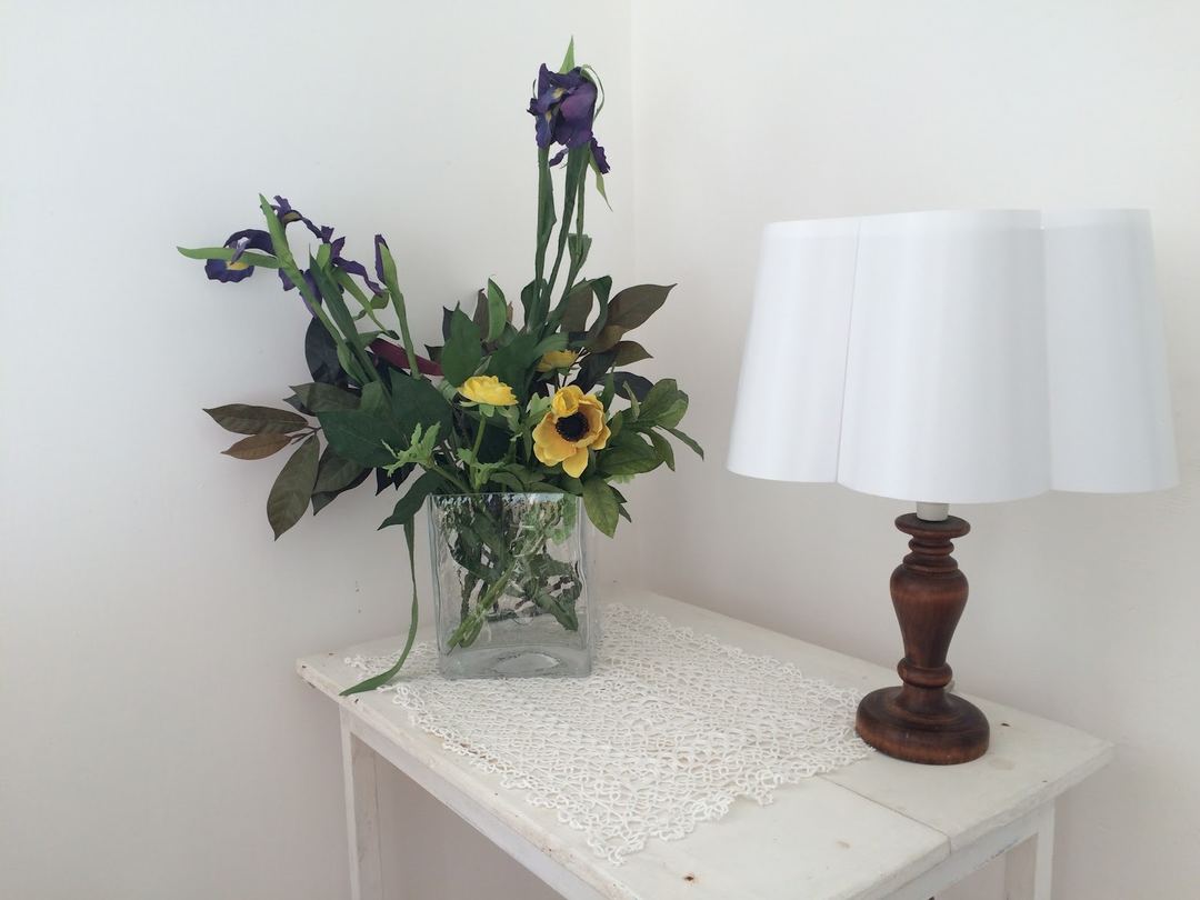 A decorative white side table displays a floral arrangement with purple irises and yellow flowers alongside a wooden table lamp with a white shade.